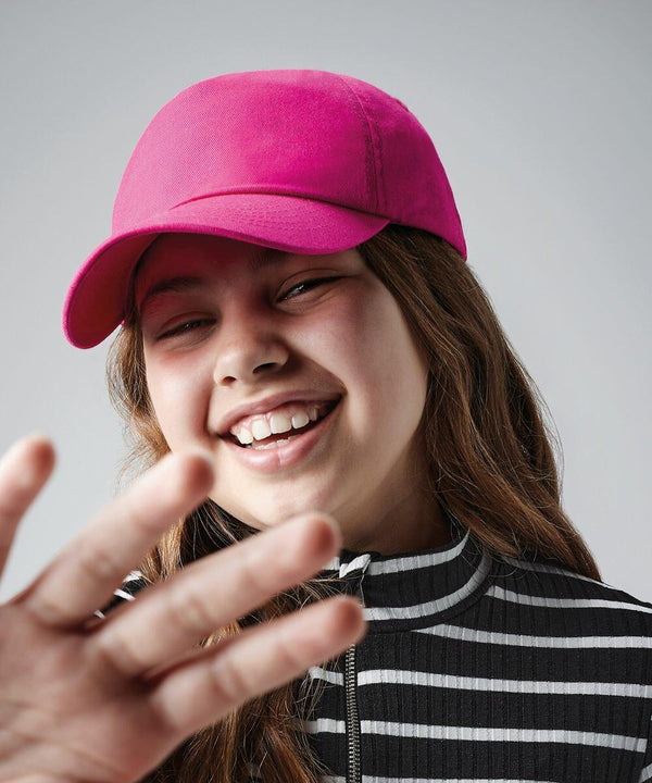 A person with long hair is smiling broadly while wearing a bright pink Beechfield BC10B baseball cap and a black and white striped jacket. Their hand is raised in front of the camera, slightly blurred, giving a friendly and playful vibe. The background is neutral gray.