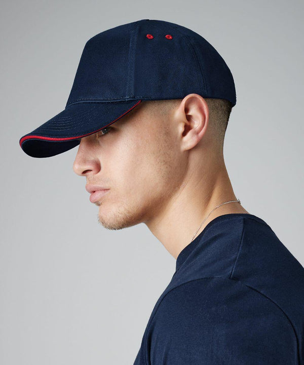 A person is shown in a profile view wearing a Beechfield BC15C navy baseball cap with red accents. They also have a dark navy shirt and a thin necklace. The background is a plain light grey gradient.