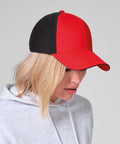 A person with light blonde hair is looking downward, wearing a Beechfield Club cap in red and black and a light grey hoodie, set against a plain light grey background.