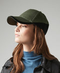A person with long brown hair is wearing a green and black Beechfield BC171, a blue ribbed turtleneck sweater, and a black jacket. They are looking slightly to the left against a plain gray background.