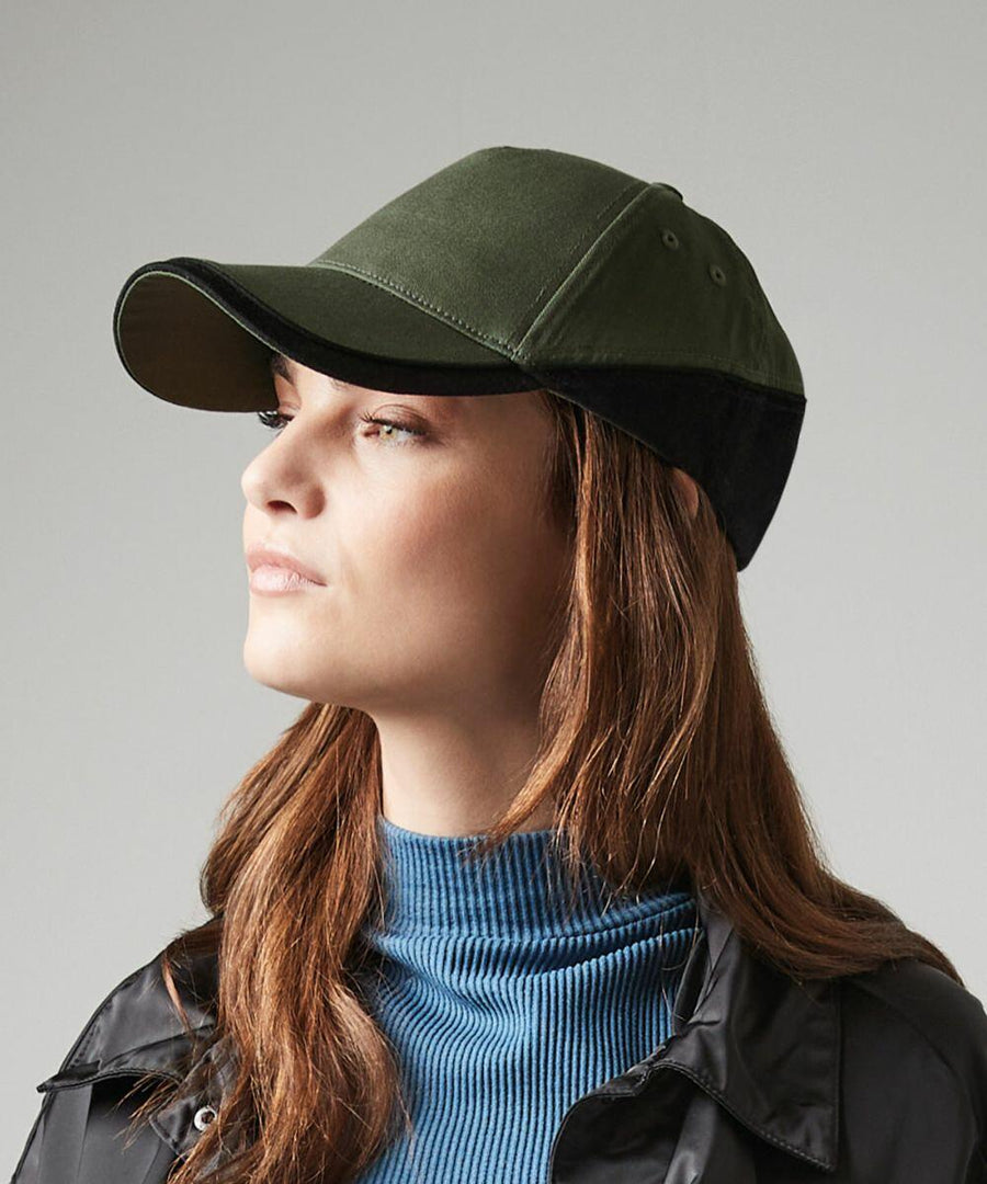 A person with long brown hair is wearing a green and black Beechfield BC171, a blue ribbed turtleneck sweater, and a black jacket. They are looking slightly to the left against a plain gray background.
