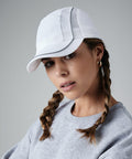 A person with two braided pigtails poses against a neutral background, wearing a white Beechfield BC182 baseball cap with mesh detailing and a light gray sweatshirt. The head is slightly tilted and there is a calm, thoughtful expression on their face.