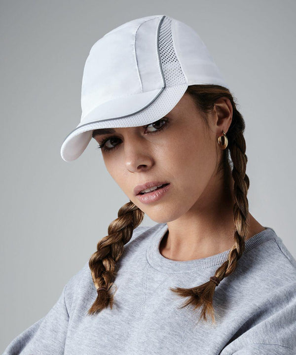 A person with two braided pigtails poses against a neutral background, wearing a white Beechfield BC182 baseball cap with mesh detailing and a light gray sweatshirt. The head is slightly tilted and there is a calm, thoughtful expression on their face.