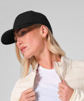 A woman wears the Beechfield Technical Training Cap, gold hoop earrings, a white shirt, and a light quilted jacket as she looks to the side against a plain background.