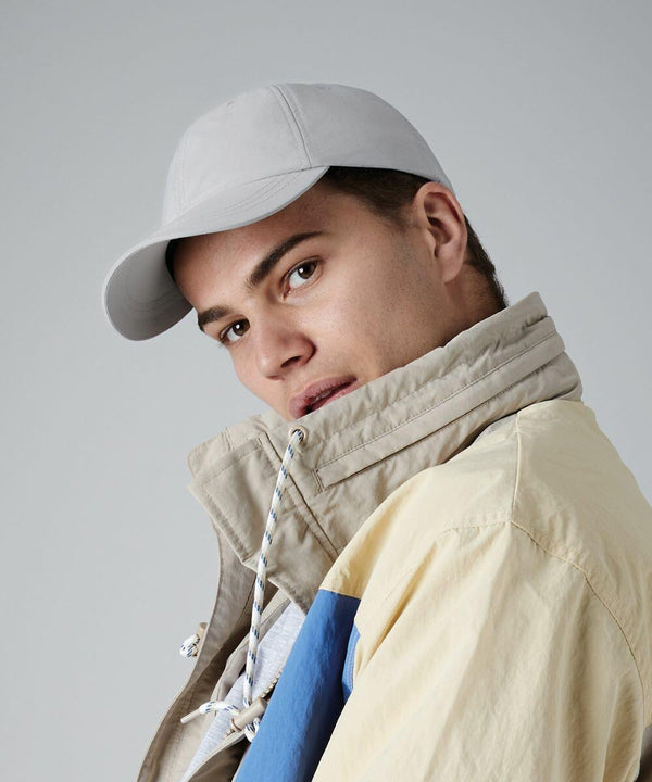 A person wearing a light gray baseball cap and a beige and blue Beechfield BC187 jacket with a high collar looks directly at the camera against a plain background. The jacket's strings are loosely hanging.
