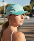 A person with long hair pulled back, wearing a light blue Beechfield BC188 baseball cap and a white tank top, looks over their shoulder. Sunlight illuminates their face. The background is an outdoor setting with blurred trees and buildings.