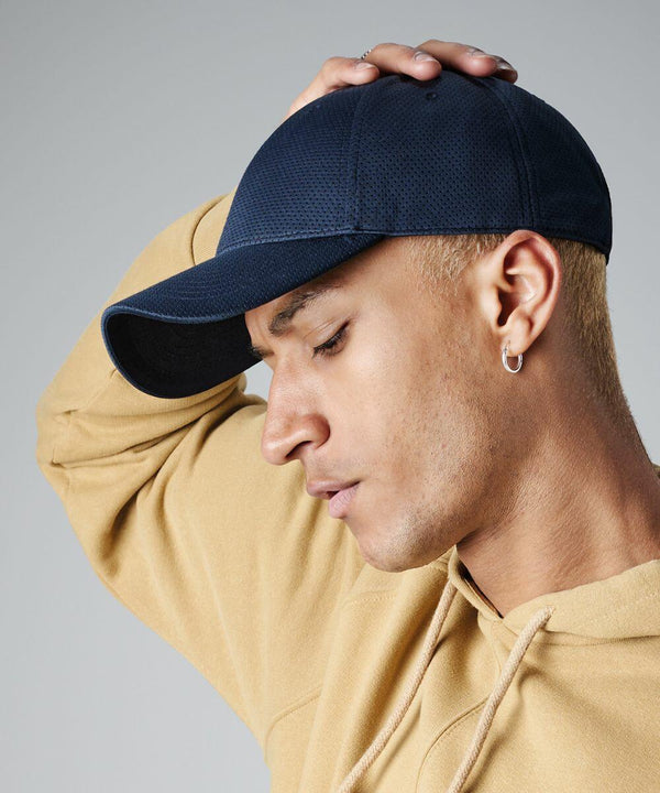 A person with short blond hair is wearing a dark blue Beechfield BC196 and a beige hoodie. They are holding the back of their head with one hand and looking downwards. They have a small hoop earring in their right ear.