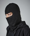 Person wearing a black Beechfield BC225 covering their head and neck, leaving only their eyes exposed. They are dressed in a black hoodie, facing to the left, with a neutral gray background.