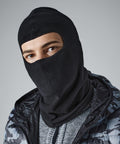 A person wearing a black Beechfield BC230 that covers most of their face, leaving only their eyes visible. They are also dressed in a dark patterned jacket with light and dark grey tones. The background is plain and light grey.