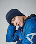 A person wearing a Beechfield BC244 blue knit beanie and a blue puffer jacket is tilting their head slightly to the side while looking at the camera against a plain gray background.