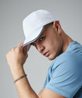 A person wearing a white Beechfield BC25C and a light blue T-shirt looks intently at the camera while slightly tilting the cap with one hand. They also wear a silver chain bracelet on their wrist. The background is plain gray.