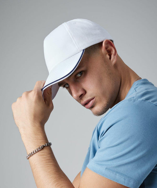 A person wearing a white Beechfield BC25C and a light blue T-shirt looks intently at the camera while slightly tilting the cap with one hand. They also wear a silver chain bracelet on their wrist. The background is plain gray.