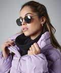 A person with long, brown hair is wearing mirrored aviator sunglasses, small hoop earrings, a black scarf, and a light purple Beechfield BC285 puffer jacket. They are holding their jacket collar with both hands and looking to the side against a gray background.