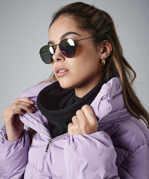 A person with long, brown hair is wearing mirrored aviator sunglasses, small hoop earrings, a black scarf, and a light purple Beechfield BC285 puffer jacket. They are holding their jacket collar with both hands and looking to the side against a gray background.