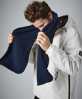 A person with curly hair is wearing a white jacket with a zippered pocket on the side and holding a navy blue Beechfield BC290 up to their face, appearing to adjust or inspect it. The background is plain and neutral.