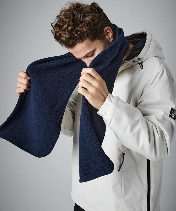 A person with curly hair is wearing a white jacket with a zippered pocket on the side and holding a navy blue Beechfield BC290 up to their face, appearing to adjust or inspect it. The background is plain and neutral.