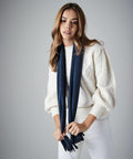 A person with long brown hair, wearing a white sweater with cable-knit detailing and white pants, holds a dark blue Beechfield BC291 fringed scarf. The individual stands against a plain, light gray backdrop.