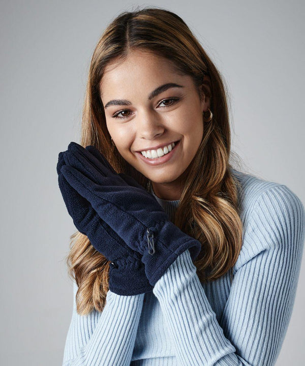 A young woman with long, wavy brown hair smiles warmly at the camera. She is wearing a light blue ribbed sweater and holding her navy blue gloved hands up to her face, appearing cozy and cheerful in Beechfield's BC295, against a plain gray background.