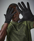 A man wearing a green athletic shirt and black Beechfield BC310 gloves holds his hands up towards the camera, partially covering his face. The gloves have textured patterns on them, and he appears to be posing in front of a neutral gray background.