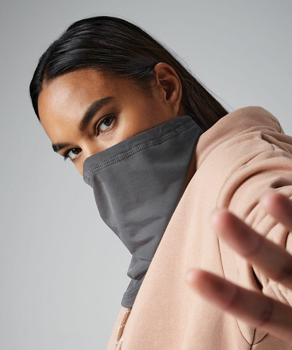 Person with long dark hair wearing a beige hoodie and a gray Beechfield BC320 face covering that covers their nose and mouth, extending a hand toward the camera, creating a sense of depth. The background is plain and softly lit, emphasizing the person's focused gaze.