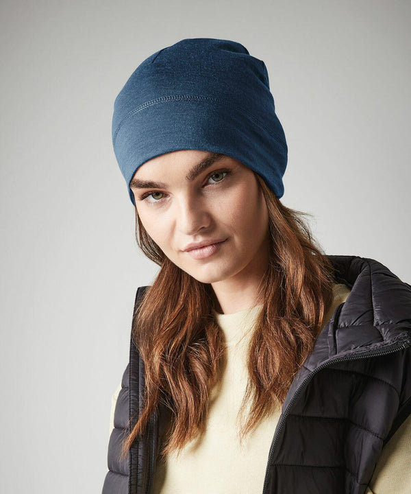 A person with long brown hair is wearing a blue Beechfield BC370 beanie, a light yellow top, and a black quilted vest. They are looking at the camera with a neutral expression. The background is plain and light-colored.