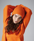 A person wearing a Beechfield BC380 orange beanie and an orange sweatshirt stretches with arms raised behind the head, one crossing over the other. They have long wavy brown hair and look into the camera with a neutral expression. The background is plain gray.