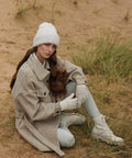 A woman with long brown hair sits on sandy ground surrounded by grass, wearing a light beige Beechfield BC387 coat, matching knit hat, gloves, white leggings, and boots. She gazes calmly at the camera, exuding a stylish and cozy vibe.