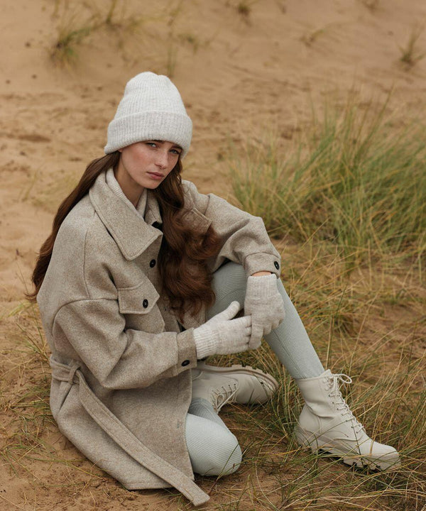 A woman with long brown hair sits on sandy ground surrounded by grass, wearing a light beige Beechfield BC387 coat, matching knit hat, gloves, white leggings, and boots. She gazes calmly at the camera, exuding a stylish and cozy vibe.