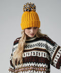 A person with long blonde hair is wearing a yellow Beechfield BC390 knit hat with a multicolored pom-pom and a patterned sweater featuring dark red, black, and white designs. The person is looking down with a neutral expression. The background is plain gray.