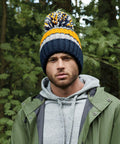 A man with a beard wearing a green rain jacket, gray hoodie, and a colorful Beechfield BC392 beanie with blue, orange, white, and yellow stripes stands in front of a green, forested background. He looks directly at the camera with a neutral expression.
