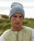 A person wearing a light gray Beechfield BC40R beanie, a beige quilted vest, and a white shirt stands outdoors. The background is blurred with green foliage, suggesting a natural setting.