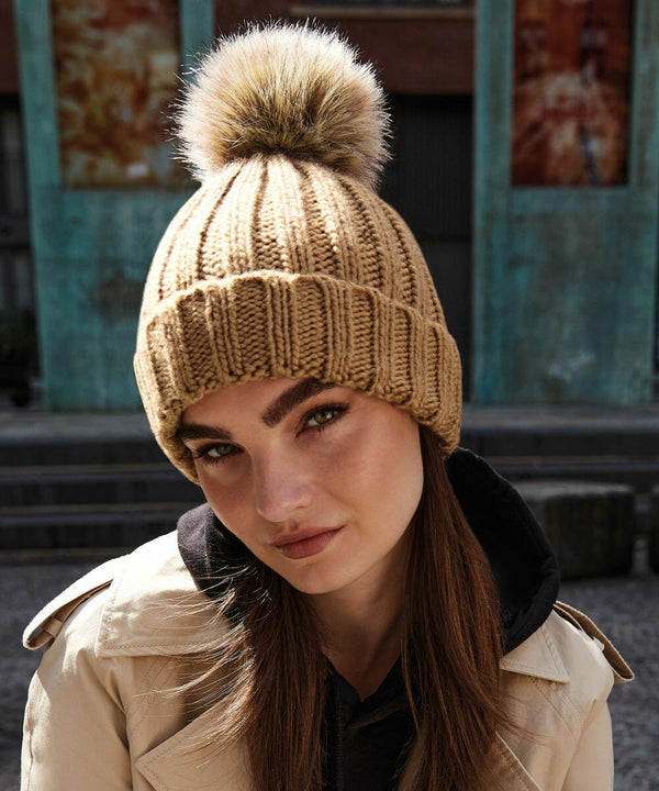 A person with long brown hair is wearing a Beechfield BC412 beanie with a large, fluffy pom-pom on top. They are dressed in a beige trench coat over a black hoodie and are standing outdoors in front of a blue-green building. The person’s expression is neutral.