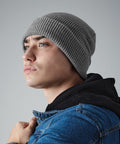 A person wearing a gray knit Beechfield BC425, a black hoodie, and a blue denim jacket looks upwards with a serious expression. The background is a plain gray.
