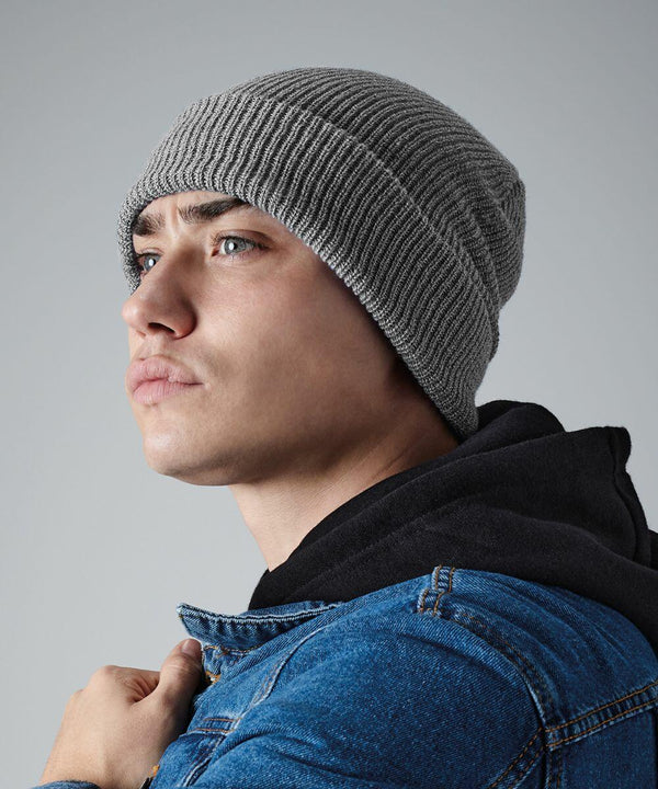 A person wearing a gray knit Beechfield BC425, a black hoodie, and a blue denim jacket looks upwards with a serious expression. The background is a plain gray.