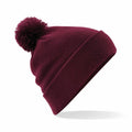 A Beechfield BC426 maroon knit beanie with a large pom-pom on top is displayed against a white background. It is slightly angled to showcase its folded cuff.