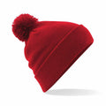 A Beechfield BC426 red knitted beanie featuring a pom-pom on top is displayed against a white background. The beanie includes a folded brim, enhancing its cozy and casual appearance.