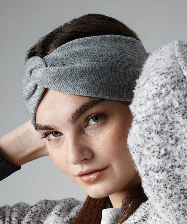 A person with long brown hair is wearing a Beechfield BC432 light gray knitted headband with a twist knot design in the front. They are dressed in a cozy, textured gray sweater and have one arm raised, partially obscuring the background, which is plain and gray.