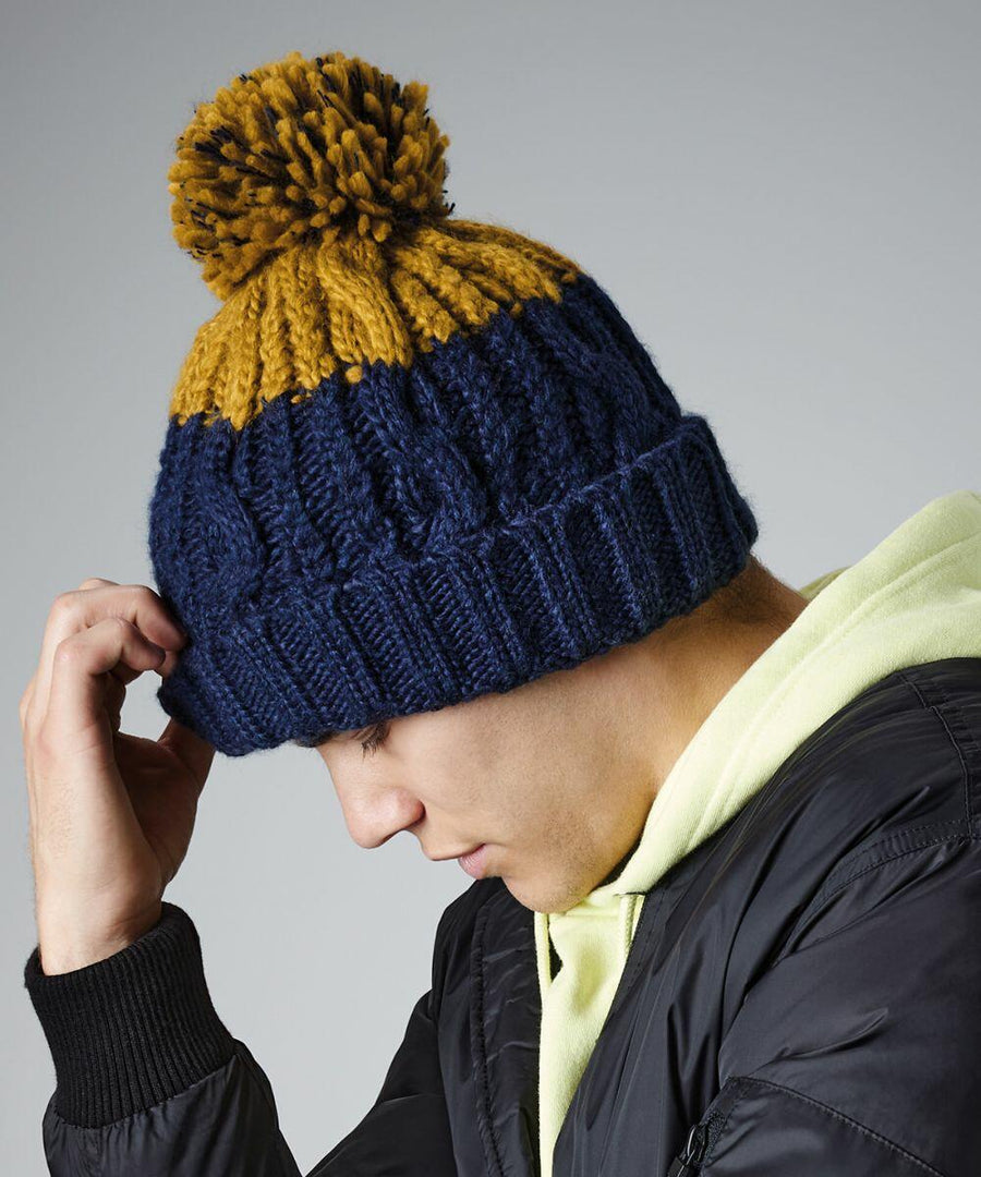 A person wearing a dark-colored jacket and a light-colored hoodie is looking down and adjusting their blue and yellow Beechfield BC437 pom-pom knit hat. The studio background is plain gray.