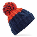 Introducing the BC437 by Beechfield: a stylish knitted beanie featuring a vibrant red-orange pom-pom. This hat showcases a two-tone design, combining an upper section in red-orange with a lower section in navy blue. It is adorned with intricate cable knit patterns and finished with a ribbed band.