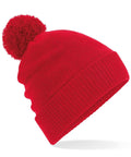 The BC439 knit beanie from Beechfield features a vibrant red color and a large pom-pom on top. Its simple design is highlighted against a white background, showcasing the hat's texture and color.