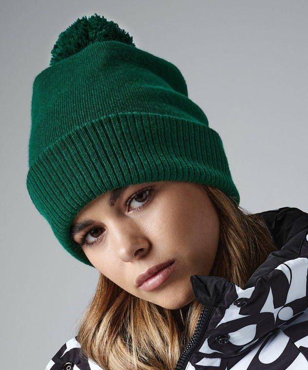 A person with long brown hair is wearing a green Beechfield BC439 knit beanie with a pom-pom. They are dressed in a black and white patterned winter jacket. The background is plain and grey, and the person is looking slightly downwards.