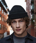 A young person with curly hair and blue eyes is wearing a black Beechfield BC43R beanie and dark jacket. They are standing in an urban environment with brick buildings blurred in the background. The image captures the person from the shoulders up.