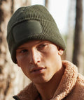 A person with a serious expression is wearing a green knit beanie from Beechfield with a BC440 patch on the front. They are in an outdoor setting with blurred trees in the background and are dressed in a tan, sherpa-lined jacket.