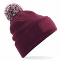 The BC443 by Beechfield is a maroon knit beanie featuring a pom-pom on top, a ribbed fold-over cuff, and a blank rectangular patch on the front.