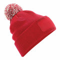 The Beechfield BC443 is a red knitted beanie adorned with a red and white pom-pom on top, featuring a ribbed band and a blank patch on the front. The design is slightly angled to highlight its texture and craftsmanship.