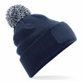 A Beechfield BC443 knit beanie in navy blue, featuring a white and navy mixed fluffy pom-pom on top. It comes with a ribbed fold-over cuff and a blank patch on the front, worn tilted slightly to the side.