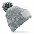The Beechfield BC443 is a light gray knit beanie featuring a pom-pom on top and a blank rectangular patch on the front, displayed against a white background.