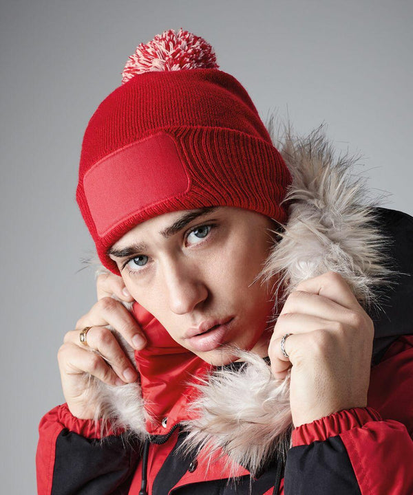 A person wearing a red Beechfield BC443 beanie with a pom-pom and a red and black winter jacket with a fur-lined hood. They are looking directly at the camera while holding the fur trim of the jacket. The background is a plain, light grey color.