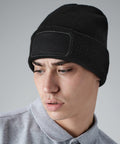 A person with short hair is wearing a black Beechfield BC445 knit beanie with a blank rectangular patch on the front. They are dressed in a gray collared shirt and are looking downwards to their left against a plain gray background.
