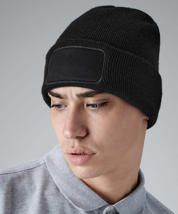 A person with short hair is wearing a black Beechfield BC445 knit beanie with a blank rectangular patch on the front. They are dressed in a gray collared shirt and are looking downwards to their left against a plain gray background.
